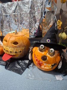 Dwie dynie w stylu halloweenowym. Pierwsza ma szeroki uśmiech z „zębami” z przypinek i wystający czerwony język. Druga dynia przedstawia czarownicę z kapeluszem, dużymi oczami z drucianych kulek i sztucznymi włosami.