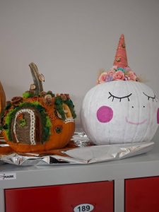 Dwie ozdobione dynie stojące na blacie. Jedna pomalowana na biało z rogiem jednorożca i kwiatami na górze. Druga dynia udekorowana jak mały domek z mchem, szyszkami i drzwiami z sznurka.