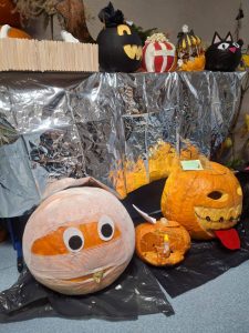 Kilka kreatywnie przystrojonych dyni ustawionych na wystawie. Dynie przedstawiają m.in. mumię, potwora z czerwonym językiem, lampion z pajęczyną oraz dynię popcorn.