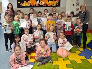 Grupa dzieci wraz z organizatorką konkursu i panią dyrektor stoi na szkolnym korytarzu. Dzieci trzymają dyplomy i nagrody za konkurs na najbardziej kreatywną jesienną dynię.