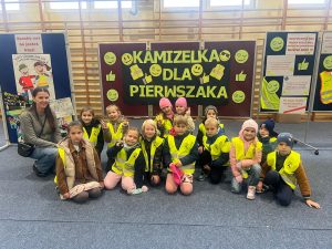 Grupa uczniów klas pierwszych w kamizelkach odblaskowych pozuje z nauczycielką przed planszą „Kamizelka dla pierwszaka”. W tle widoczne są kolorowe plakaty przypominające o zasadach bezpieczeństwa na drodze.