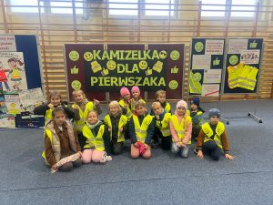 Pierwszoklasiści w kamizelkach odblaskowych siedzą i klęczą przed planszą z napisem „Kamizelka dla pierwszaka”. Dzieci pozują do wspólnego zdjęcia w sali gimnastycznej, za nimi widoczne są tablice edukacyjne i kamizelki odblaskowe.