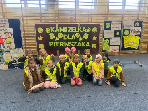 Pierwszoklasiści w kamizelkach odblaskowych siedzą i klęczą przed planszą z napisem „Kamizelka dla pierwszaka”. Dzieci pozują do wspólnego zdjęcia w sali gimnastycznej, za nimi widoczne są tablice edukacyjne i kamizelki odblaskowe.