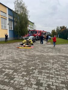 Na placu przed szkołą odbywa się symulacja wypadku drogowego. Strażacy udzielają pomocy osobie poszkodowanej, w tle stoją uczniowie i nauczyciele obserwujący działania służb.