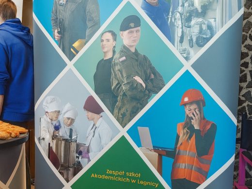 Baner promocyjny Zespołu Szkół Akademickich w Legnicy złożony z sześciu zdjęć przedstawiających uczniów w różnych zawodach, m.in. kucharzy, żołnierza, technika, budowlańca i informatyka. Baner stoi przy ścianie o mozaikowym wzorze, obok stolika z przekąskami.