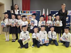 Grupa pierwszoklasistów w szkolnych togach i biretach pozuje do zdjęcia z nauczycielkami. Dzieci trzymają w rękach zestawy upominkowe z dyplomami. W tle widoczny jest szkolny plakat z napisem „Różni, ale tacy sami…” oraz polska flaga.