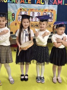 Cztery dziewczynki w galowych strojach i granatowych biretach stoją w szeregu przed kolorową dekoracją z napisem „Różni, ale tacy sami...”. Trzy z nich mają ręce skrzyżowane na piersiach, a jedna trzyma tamburyn. W tle widać plakat z ilustracją szkolnego autobusu i zdjęciami dzieci.