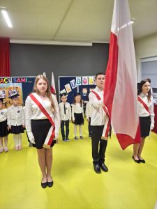 Poczet flagowy wprowadza flagę Polski podczas uroczystości szkolnej. Troje starszych uczniów w białych koszulach i z biało-czerwonymi szarfami stoi na przodzie sali. Za nimi ustawieni są pierwszoklasiści w biretach, gotowi do pasowania na ucznia. W tle widać dekoracje z napisami „Różni, ale tacy sami” i „Ślubowanie”.