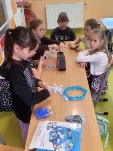 Zdjęcie 10 (p11.jpg) Grupa uczennic siedzi przy szkolnym stole i gra w gry planszowe oraz karciane. Na blacie widać elementy kilku gier, w tym kolorowe klocki i planszę. Dzieci są skupione na wspólnej zabawie.