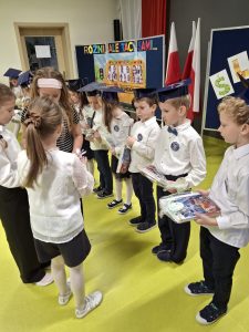 Dzieci ubrane w białe koszule i granatowe birety stoją w szeregu podczas uroczystości pasowania na ucznia. Każde dziecko trzyma w rękach upominek z dyplomem.