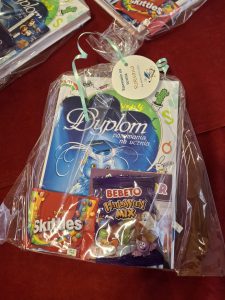 Zestaw upominkowy dla ucznia zapakowany w przezroczystą folię, zawierający dyplom „Pasowania na ucznia”, paczkę cukierków Skittles i żelki Bebeto Halloween Mix. Na zestawie widać zawieszkę z imieniem „Roksana”.