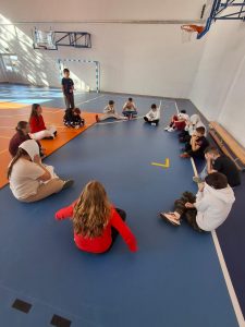 Grupa uczniów siedzi w kole na sali gimnastycznej. Dzieci uczestniczą w zajęciach integracyjnych, rozmawiają i słuchają swojego kolegi stojącego za nimi.