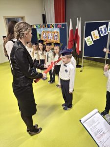 Chłopiec w białej koszuli i granatowym birecie zostaje pasowany na ucznia przez panią wicedyrektor, która dotyka jego ramienia dużym czerwonym ołówkiem. W tle widać inne dzieci ustawione w rzędzie oraz dekorację z napisem „Różni, ale tacy sami...” i polskie flagi.