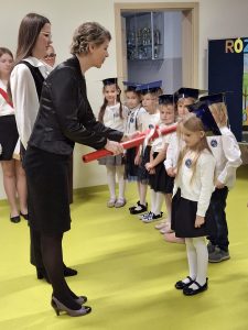 Pani wicedyrektor pasuje na ucznia dziewczynkę w białym sweterku i czarnej spódniczce, trzymając duży czerwony ołówek. Obok stoją inne dzieci w białych koszulach i biretach, czekające na swoją kolej. W tle widoczna jest tablica z napisem „Różni, ale tacy sami...”.