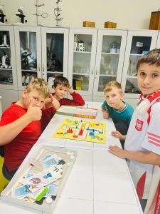 Czterech uczniów siedzi przy stole i gra w grę planszową „Chińczyk”. Na twarzach widać uśmiechy, a w tle znajdują się szkolne szafki i sprzęt laboratoryjny.