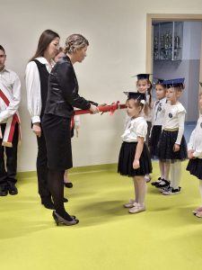 Pani wicedyrektor ubrana na czarno dokonuje pasowania na ucznia dziewczynki w białej bluzce i czarnej spódniczce, dotykając jej ramienia dużym, czerwonym ołówkiem. Obok stoi wychowawczyni klasy, a w tle widać inne dzieci w granatowych biretach, czekające na swoją kolej.