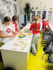 Uczniowie pracują w grupach przy stołach w sali lekcyjnej. Grają w różne gry planszowe i karciane. W tle widoczne są szkolne meble i pomoce naukowe.