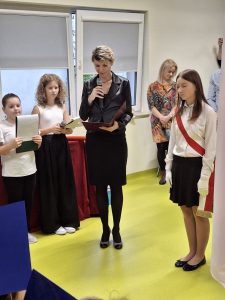 Wicedyrektor szkoły ubrana na czarno przemawia do uczniów, trzymając mikrofon i teczkę z dokumentami. Obok stoją uczniowie w galowych strojach – jedna dziewczynka z biało-czerwoną szarfą oraz dwoje dzieci z kartkami w rękach. W tle widoczni są dorośli obserwujący uroczystość.