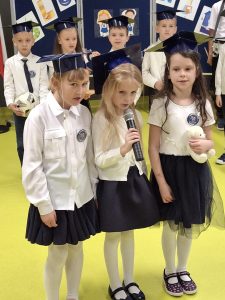 Trzy dziewczynki w szkolnych strojach galowych i granatowych biretach stoją razem, śpiewając lub recytując do mikrofonu. Za nimi widać innych uczniów pierwszej klasy w białych koszulach i biretach, stojących na tle szkolnej dekoracji.