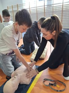 Instruktorka pokazuje uczniowi, jak prawidłowo wykonywać uciski klatki piersiowej na fantomie podczas zajęć z pierwszej pomocy w sali gimnastycznej. W tle inni uczniowie ćwiczą na swoich fantomach.