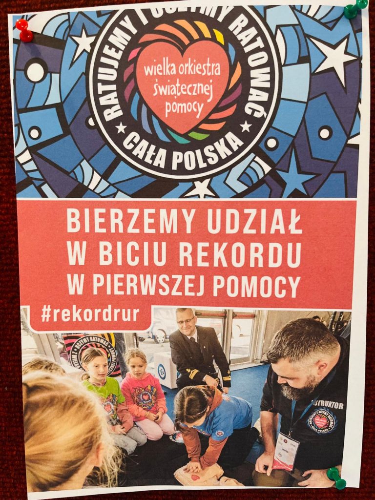 Plakat z logo Wielkiej Orkiestry Świątecznej Pomocy i napisem: ‘Ratujemy i uczymy ratować. Cała Polska. Bierzemy udział w biciu rekordu w pierwszej pomocy. #rekordrur’. Na dole zdjęcie dzieci uczących się resuscytacji pod okiem instruktora.