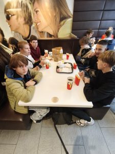 Uczniowie siedzący przy stolikach w restauracji McDonald’s, jedzący frytki i burgery. Na stole widać torbę z jedzeniem i napoje w charakterystycznych kubkach.