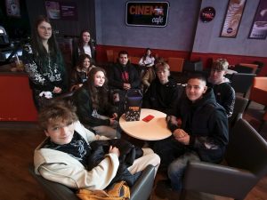 Grupa młodzieży w kawiarni Cinema Café, siedząca przy stolikach przed seansem filmowym. W tle widoczne logo kawiarni i plakaty napojów.