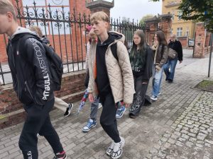 Grupa uczniów idąca chodnikiem wzdłuż ceglanego ogrodzenia, w jesiennych ubraniach. W tle widać budynki i liście na drzewach.