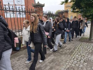 Uczniowie idący ulicą wzdłuż ceglanego ogrodzenia, ubrani w ciepłe kurtki. W tle widać budynki miasta i drzewa z jesiennymi liśćmi.