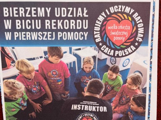 Plakat akcji ‘Ratujemy i uczymy ratować’ z logo Wielkiej Orkiestry Świątecznej Pomocy. Widoczne dzieci w kolorowych koszulkach z logo WOŚP uczą się udzielania pierwszej pomocy pod opieką instruktora.