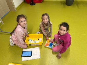 TTrzy dziewczynki siedzą na zielonej podłodze w sali. Bawią się zestawem klocków LEGO Education w żółtym pudełku. Obok nich leży tablet z uruchomioną aplikacją sterującą. Wszystkie patrzą w stronę aparatu i uśmiechają się.