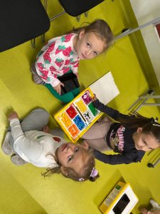 Trzy małe dziewczynki siedzą na podłodze w sali. Przed nimi znajduje się żółte pudełko z kolorowymi klockami edukacyjnymi LEGO. Jedna dziewczynka trzyma tablet, pozostałe oglądają klocki.