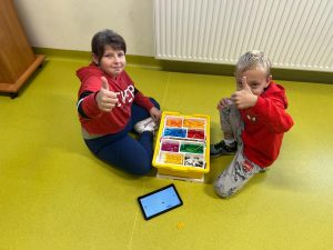 Dwójka dzieci siedzi na podłodze w sali. Przed nimi znajduje się żółte pudełko z posegregowanymi kolorowymi klockami LEGO. Oboje pokazują kciuki w górę i uśmiechają się. Na podłodze obok leży tablet.