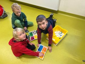 Trzech chłopców siedzi na podłodze w sali. Przed nimi żółte pudełko z posegregowanymi kolorowymi klockami LEGO. Jeden z chłopców wskazuje na klocki, drugi leży oparty na rękach, a trzeci patrzy w ich stronę.