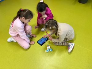 Trzy dziewczynki siedzą na podłodze w sali. Dwie z nich obserwują tablet, a jedna dotyka ekranu, wpisując komendy. Obok tabletu znajduje się robot zbudowany z klocków LEGO.
