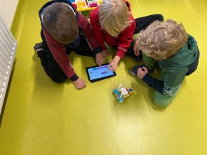 Trzech chłopców siedzi na podłodze w sali. Skupiają się na tablecie, na którym działa aplikacja do programowania. Obok stoi mały robot zbudowany z klocków LEGO na kółkach.