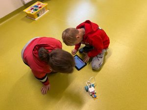 Dwójka dzieci siedzi na podłodze w sali. Skupiają się na tablecie, na którym działa aplikacja do programowania. Obok nich stoi mały robot zbudowany z klocków LEGO na kółkach. W tle widoczne jest żółte pudełko z posegregowanymi klockami.