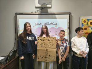 Czworo uczniów stoi przed tablicą multimedialną, na której widoczny jest napis „Siła relacji”. Uczniowie prezentują plakat z narysowanym drzewem i napisami. Obok nich stoją kolejni uczestnicy zajęć.