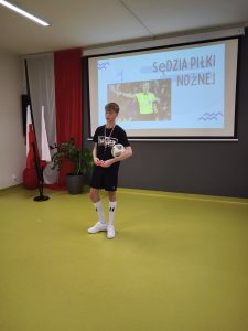 Uczeń w stroju sportowym stoi na tle prezentacji z napisem „Sędzia piłki nożnej”. W rękach trzyma piłkę. W tle widać ekran projekcyjny z napisem "sędzia piłki nożnej" oraz polską flagę.