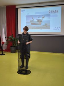 Uczeń przebrany za rybaka prezentuje zawód przed klasą. Stoi na tle slajdu z napisem „Rybak” i zdjęciem łodzi rybackiej. W rękach trzyma siatkę wędkarską.