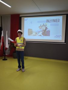 Uczeń ubrany w kask i kamizelkę odblaskową przedstawia zawód inżyniera. Za nim na ekranie widoczny jest slajd z napisem „Inżynier” oraz zdjęciem osób pracujących przy komputerach.