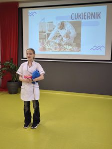 Uczennica w stroju cukiernika prezentuje zawód przed klasą. Na ekranie za nią widoczny jest napis „Cukiernik” i zdjęcie cukiernika dekorującego deser.