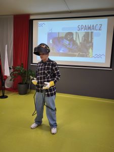 Uczeń prezentuje zawód spawacza, ubrany w kask ochronny, rękawice i koszulę w kratę. W tle widoczny jest slajd z napisem „Spawacz” i zdjęciem osoby spawającej metal.