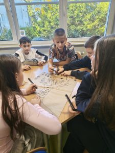 Grupa uczniów siedzi przy stole w sali lekcyjnej i wspólnie rysuje drzewo na dużym arkuszu papieru. Dzieci piszą na gałęziach słowa związane z relacjami. W tle widać duże okno i zieleń za oknem.