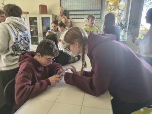 Grupa uczniów pracuje w sali biologicznej. Na pierwszym planie dwoje uczniów w bordowych bluzach korzysta ze smartfonów, a w tle pozostali uczestnicy wykonują zadania. Na półkach widać modele anatomiczne i szkielety.