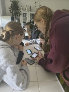 Kilkoro uczniów siedzi przy stole w sali biologicznej, korzystając ze smartfonów. Na ekranach widać aplikację edukacyjną. W tle znajdują się mikroskopy i szafki laboratoryjne.