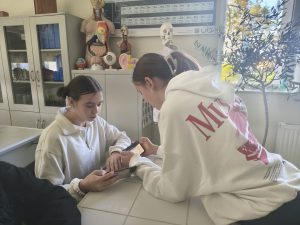 Dwie uczennice w białych bluzach pracują razem przy stole w sali biologicznej. Używają telefonów do wykonania zadania. W tle widoczne są modele anatomiczne i tablica z układem okresowym pierwiastków.