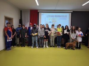 Grupa uczniów stoi wspólnie na tle prezentacji z napisem „Lekarz dermatolog”. Każdy z nich przebrany jest za przedstawiciela różnych zawodów, takich jak lekarz, budowlaniec, sportowiec czy weterynarz.