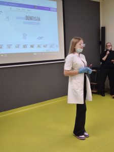 Uczennica w stroju dentystki prezentuje zawód przed klasą. Na ekranie za nią widoczny jest napis „Dentysta”. Obok stoi nauczycielka z mikrofonem.