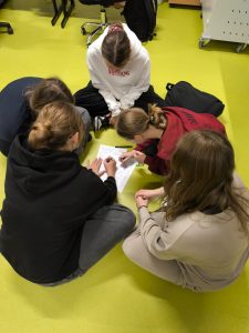 Grupa uczennic siedzi na podłodze w klasie, pracując wspólnie nad kartką z zadaniem. Jedna z nich pisze, pozostałe obserwują i dyskutują.
